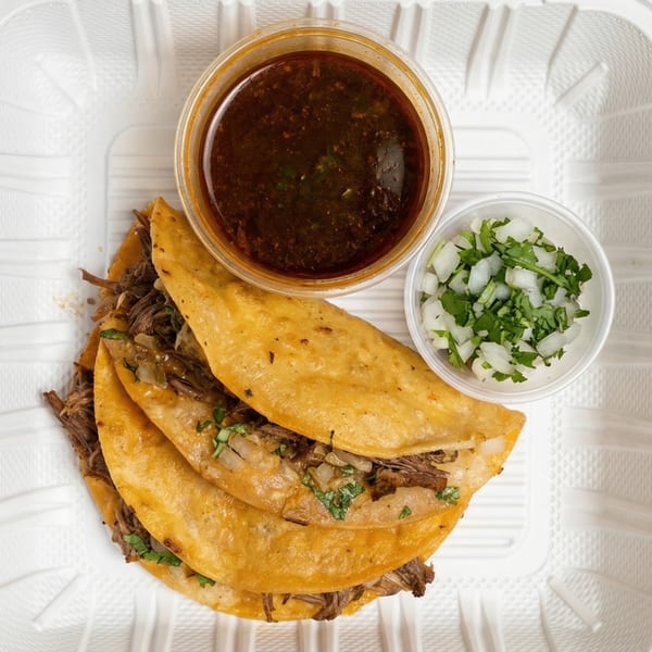 Three birria tacos with a side of consommé and a container of diced onions and cilantro.