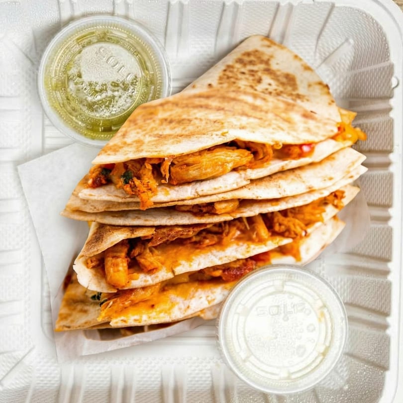 Chicken quesadillas with green and white dipping sauces in a white takeout container.