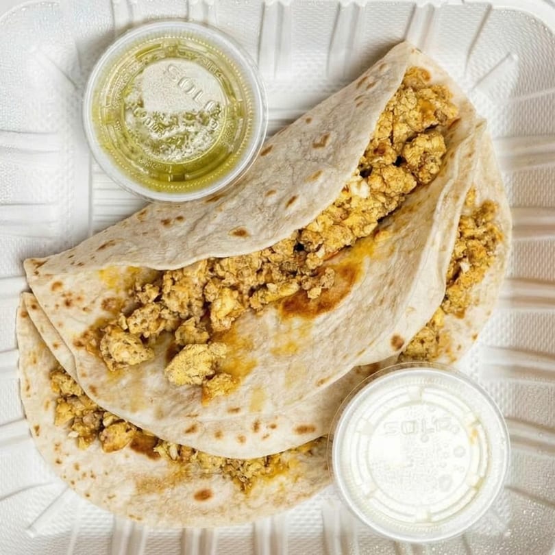 Two breakfast tacos with scrambled eggs and two small containers of salsa in a white takeout box.